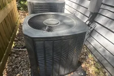 Thumbnail for Two outdoor air conditioning units on a bed of rocks next to a house and wooden fence in sunlight.