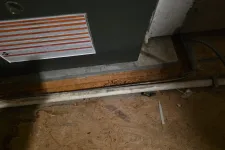 Thumbnail for Close-up of a furnace base with warning label and a white pipe on a wooden floor in a dimly lit utility area.