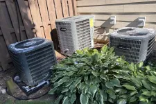 Thumbnail for Three outdoor air conditioning units behind a house next to green leafy plants and wooden fence on a sunny day