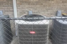 Thumbnail for Three HVAC units behind a chain-link fence against a brick wall with numbered electrical boxes above.