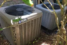 Thumbnail for Two York air conditioning units outside near a brick wall with leaves and branches around them.