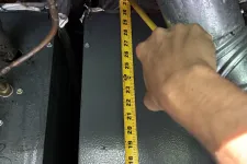 Thumbnail for Hand measuring length of metal panel with a yellow tape measure under ductwork in an industrial setting.