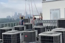 Thumbnail for Technicians installing HVAC units on a rooftop with city skyline under a cloudy sky.