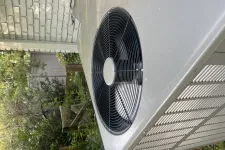 Thumbnail for Outdoor air conditioning unit with large black fan and metal casing installed beside a brick wall.