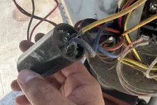 Thumbnail for Hand holding a black electrical capacitor with exposed and connected multicolored wires in a mechanical unit.
