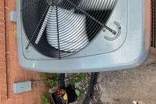 Thumbnail for Outdoor air conditioning unit with protective fan grill near brick wall and electrical equipment on concrete