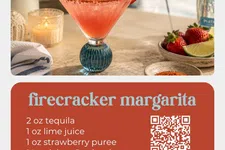 Thumbnail for Firecracker margarita cocktail with strawberry slice garnish in a blue-stemmed glass on table with ingredients and candle.