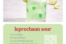 Thumbnail for Green Leprechaun Sour cocktail with lime wheel and cherry garnish in clover printed glass on counter.