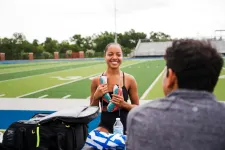 Thumbnail for Smiling female athlete holding cleats on a sports field with a coach sitting nearby during a training session