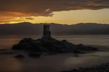 Thumbnail for Small chapel with cross on rocky islet at sunset with mountains and calm water under dramatic sky.
