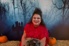 Thumbnail for Woman in red costume poses with dog dressed as Beetlejuice for Halloween near pumpkins and hay bales.
