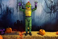 Thumbnail for Child in colorful dinosaur costume poses with hands up on hay bale stage decorated with pumpkins and Halloween lights.