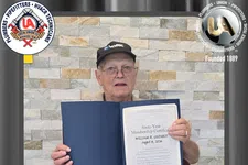 Thumbnail for Older man holding a framed sixtieth-year membership certificate seated in front of stone wall background.