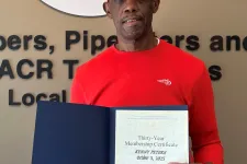 Thumbnail for Man in red shirt and cap holding a certification in front of a plumbers and service technicians union sign.