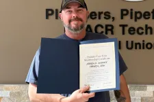 Thumbnail for Man in cap holding certificate standing by Plumbers Pipefitters Service Technicians Union logo wall