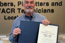 Thumbnail for Man holding a certificate standing next to Plumbers and Pipefitters Local Union 72 logo on wall