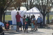 Thumbnail for Adults and children gathered outdoors near tents, engaging in a fishing activity on a sunny day with trees around.