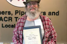 Thumbnail for Man in a red plaid shirt holding a certificate standing next to a Plumbers service union sign on a wall.