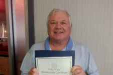 Thumbnail for Man holding a fifty-year membership certificate with a smile, standing indoors against a neutral wall.