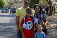 Thumbnail for Smiling family of four posing outdoors on a sunny day with trees and a small train in the background.