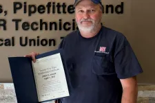 Thumbnail for Man standing next to Plumbers and Pipefitters union sign holding a certificate inside a building.