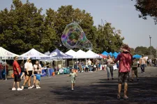 Thumbnail for Children and families enjoy a sunny outdoor market with large soap bubbles and vendor tents in the background.
