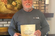 Thumbnail for Smiling man holding a certificate standing by a brick fireplace with floral painting and plants.