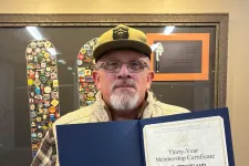 Thumbnail for Man with beard and glasses holding a thirty-year membership certificate in front of a framed pin collection.