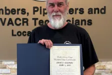 Thumbnail for Man with gray beard holding a certificate standing in front of a Plumbers and Pipefitters union sign and wall