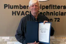 Thumbnail for Man holding certificate standing in front of Plumbers and Pipefitters Local Union 72 sign in office