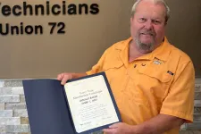 Thumbnail for Man holding certificate standing next to Plumbers, Pipefitters and HVACR Technicians Local Union 72 sign indoors.
