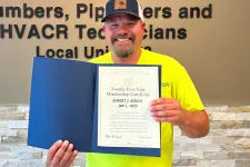 Thumbnail for Smiling man in yellow shirt holding a certificate standing beside a Plumbers and Pipefitters union sign on wall.