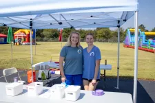 Thumbnail for Two women smiling under a white canopy tent at an outdoor event with inflatable attractions in the background