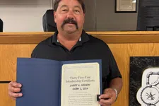 Thumbnail for Man in black shirt and shorts holding a certificate in a formal office with wooden paneling and an American flag.
