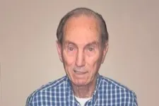 Thumbnail for Elderly man in blue plaid shirt holding a certificate and a small black box, standing indoors on wood floor.