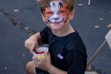 Thumbnail for Young boy with tiger face paint sitting on a chair holding a cup of snacks outdoors at a community event.