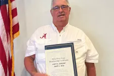 Thumbnail for Man in white shirt holding a certificate and medal standing next to an American flag indoors.