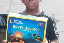 Thumbnail for Young boy holding a National Geographic Earth Science activity kit standing outdoors on a paved area.