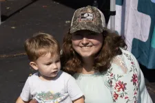 Thumbnail for Smiling woman with floral blouse and camo hat kneels next to toddler holding red paper and phone outside on pavement.