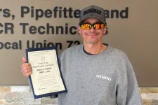 Thumbnail for Man wearing sunglasses and a Siemens sweatshirt holding a certificate next to a Plumbing and HVAC union sign.