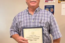 Thumbnail for Man in plaid shirt and cap holding a framed certificate standing in a room with posters on the wall.
