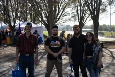 Thumbnail for Four friends standing outdoors at a sunny event with tents and trees in the background, holding blue bags.