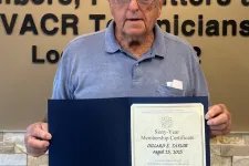 Thumbnail for Elderly man holding a certificate standing next to a Plumbers and Pipefitters union emblem on a beige wall.