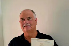 Thumbnail for Man holding a Twenty-Five-Year Membership Certificate and a lapel pin in front of a plain wall.