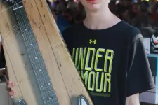Thumbnail for Boy holding boxed electric guitar at an outdoor event with tent and crowd in the background
