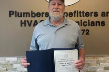 Thumbnail for Man holding a certificate standing next to Plumbers and Pipefitters Local Union 72 sign on wall.