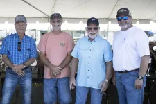 Thumbnail for Five men wearing casual clothes and caps smiling under a white event tent on a sunny day.