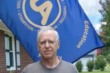 Thumbnail for Man holding a Forty-Five-Year Membership Certificate outdoors with a blue flag in the background