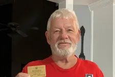 Thumbnail for Older man with white beard wearing a red Trump 2020 shirt holding a card and a small medal in a kitchen.