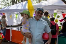 Thumbnail for Smiling man holding a toddler in a colorful hat at an outdoor market with tents and festive decorations.